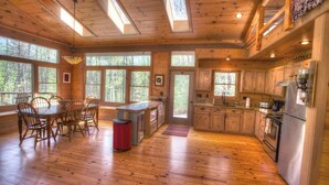 Una cabaña de madera espaciosa con comedor, cocina y grandes ventanales con vistas al bosque.
