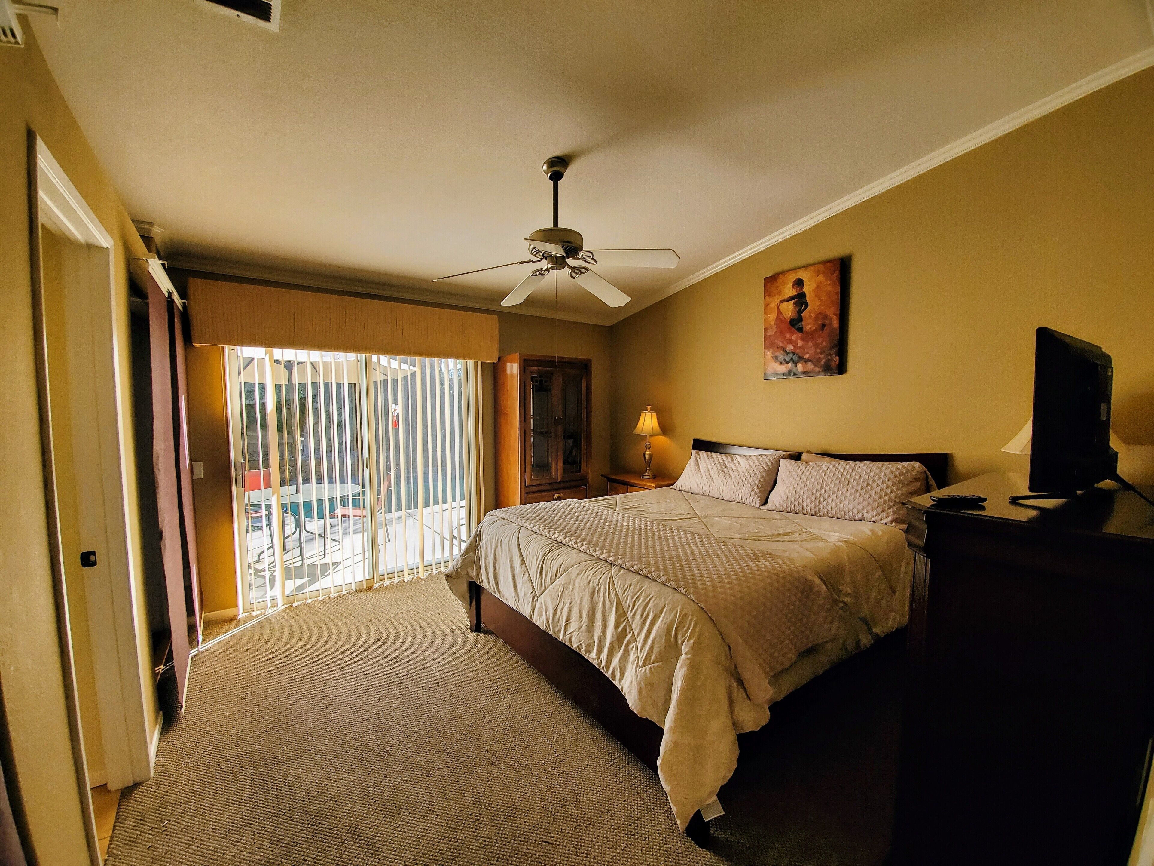 A hotel room with a bed, a ceiling fan, a TV, and a painting on the wall.