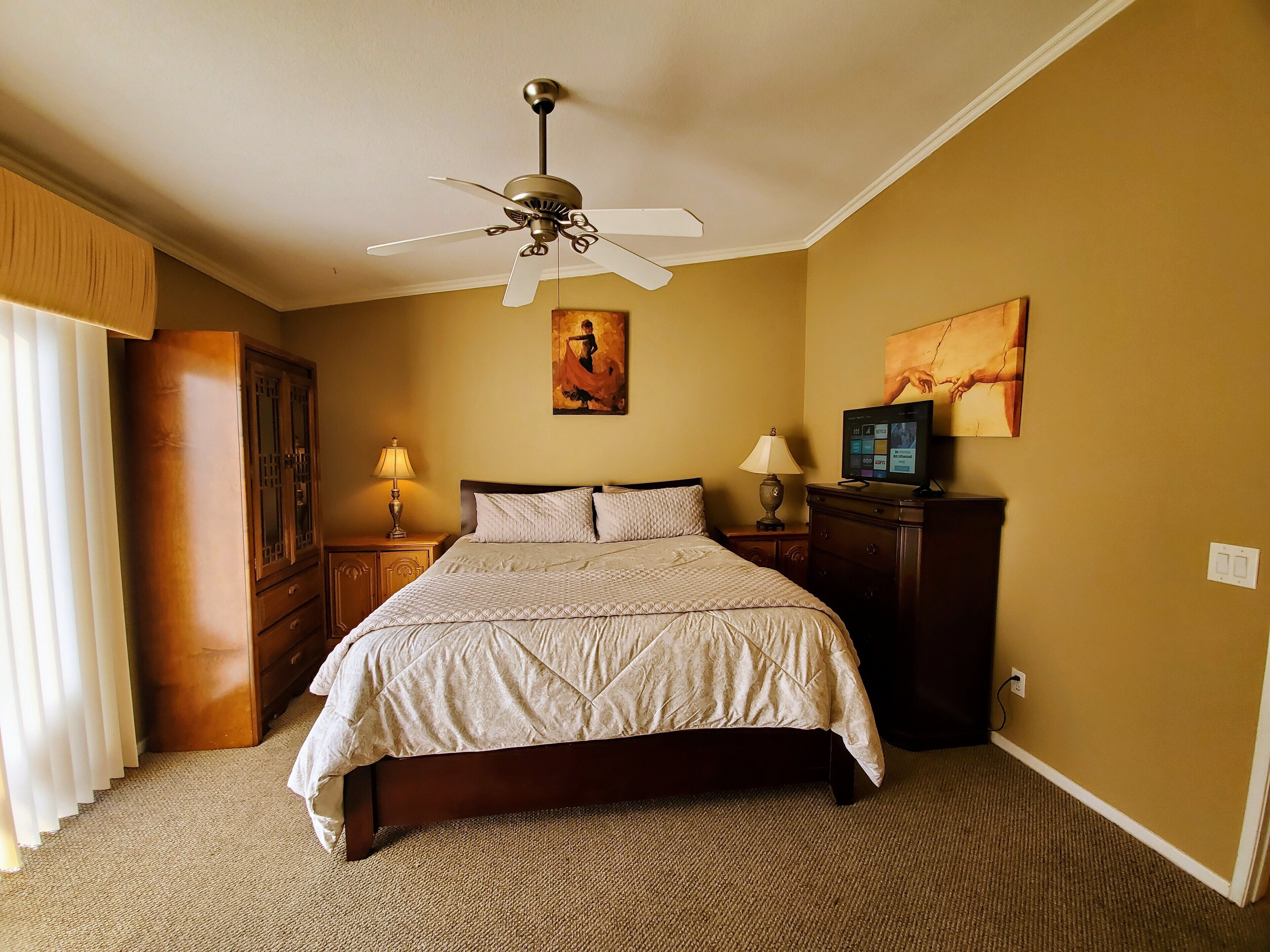 A hotel room with a bed, a ceiling fan, a television, and artwork on the walls.