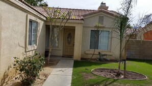 Uma casa residencial com telhado de telhas, porta da frente e um pequeno jardim com uma árvore e arbustos.