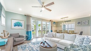 A spacious living room with a large sofa, a coffee table, and a ceiling fan. There is a dining area with a table and chairs visible through the glass doors. - 122 Luxury Hilton Head Beach House w/ Heated Pool & Elevator (Hilton Head Island)