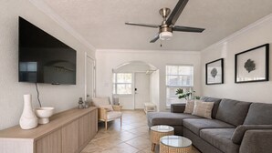 A modern living room with a grey sofa, a wooden console table, a TV mounted on the wall, and a ceiling fan. - Modern Barefoot Beach 2BR Condo, Steps to Beach & Pool (Indian Shores)