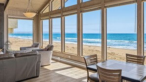 Quarto com grandes janelas dando para uma praia, mobiliado com sofá, cadeiras e uma mesa.