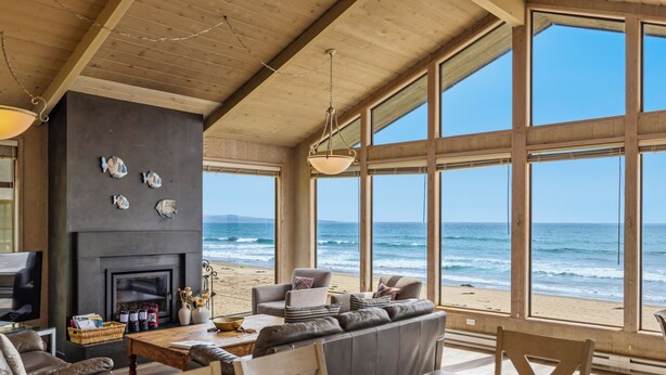 Uma sala de estar ampla com uma grande janela com vista para a praia, lareira e um espaço de estar acolhedor.