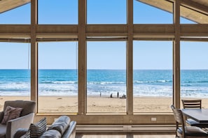 Blick auf einen Strand von einem Zimmer aus, mit großen Fenstern, Holzbalken und einer Sitzecke.