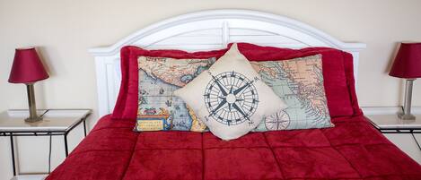 A bed with a red quilt and two decorative pillows, one with a compass design and another with a map pattern.