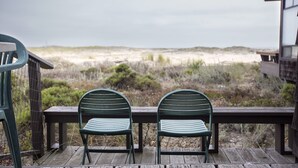 Twee groene metalen stoelen op een houten terras met uitzicht op een strand met duinen en schaarse begroeiing.