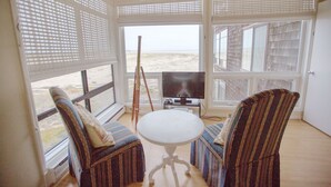 Een kamer met twee gestoffeerde fauteuils, een kleine ronde tafel, een televisie en een schilderij op een driepoot. Grote ramen met zonwering bieden uitzicht op het strand.