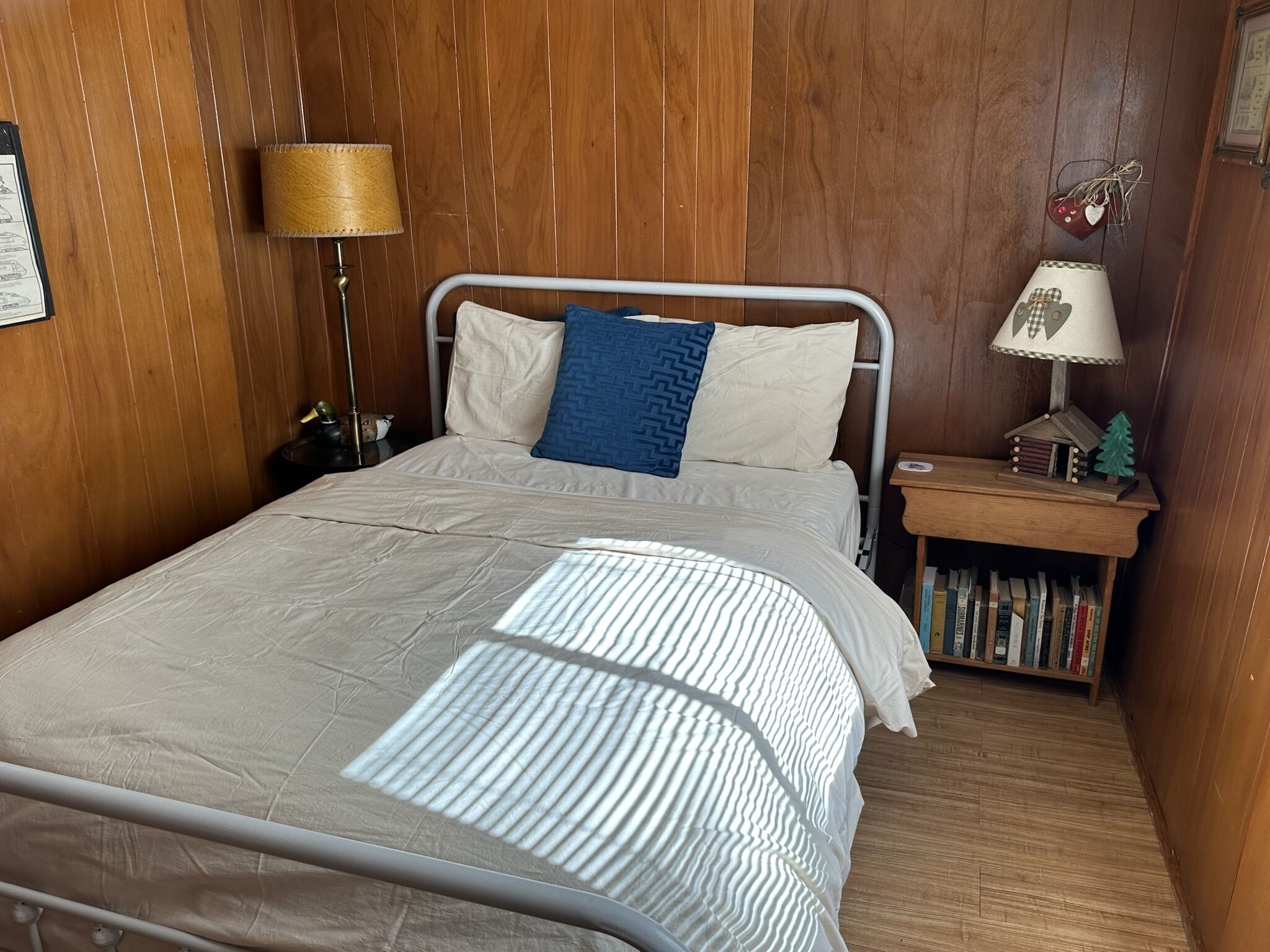 A neatly made bed with a striped blanket, a wooden nightstand with a lamp, and a small bookshelf with books and a decorative item.