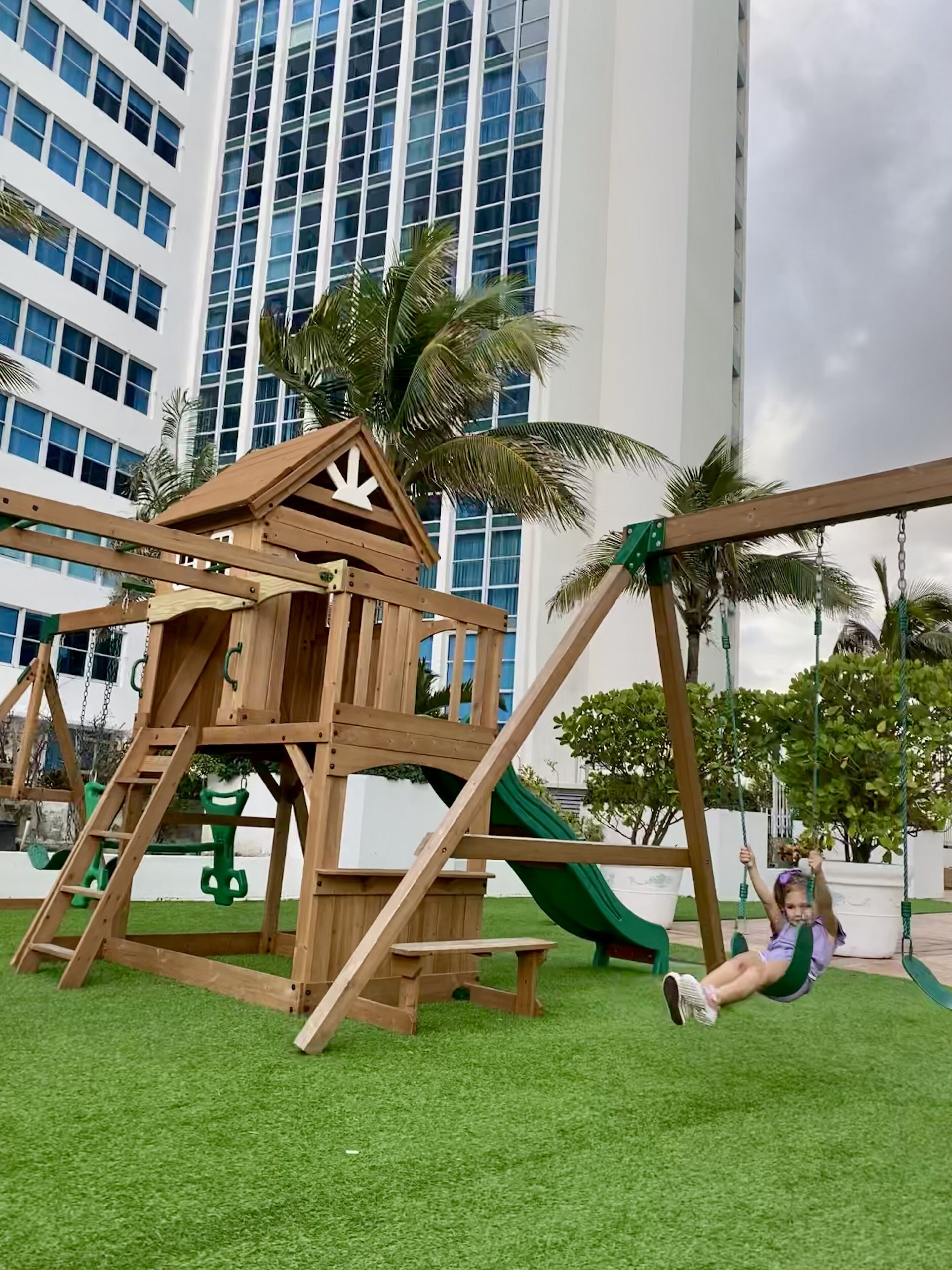 A wooden playground with a slide, swings, and a climbing structure, set against a backdrop of a modern building and palm trees.