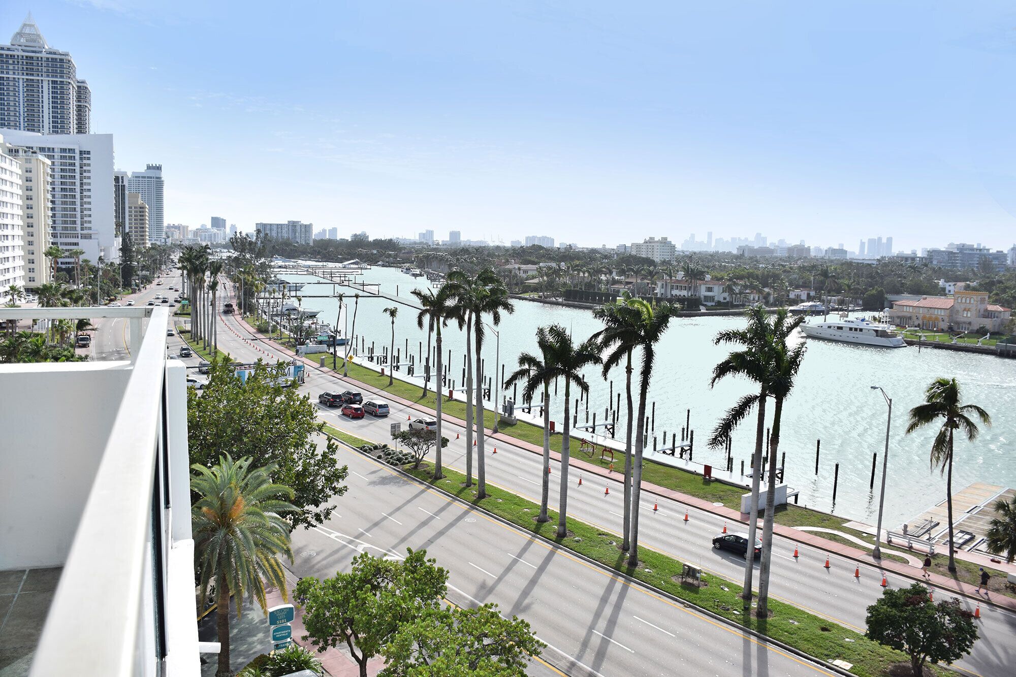 A view of a waterfront road lined with palm trees, a marina, and high-rise buildings in the background.