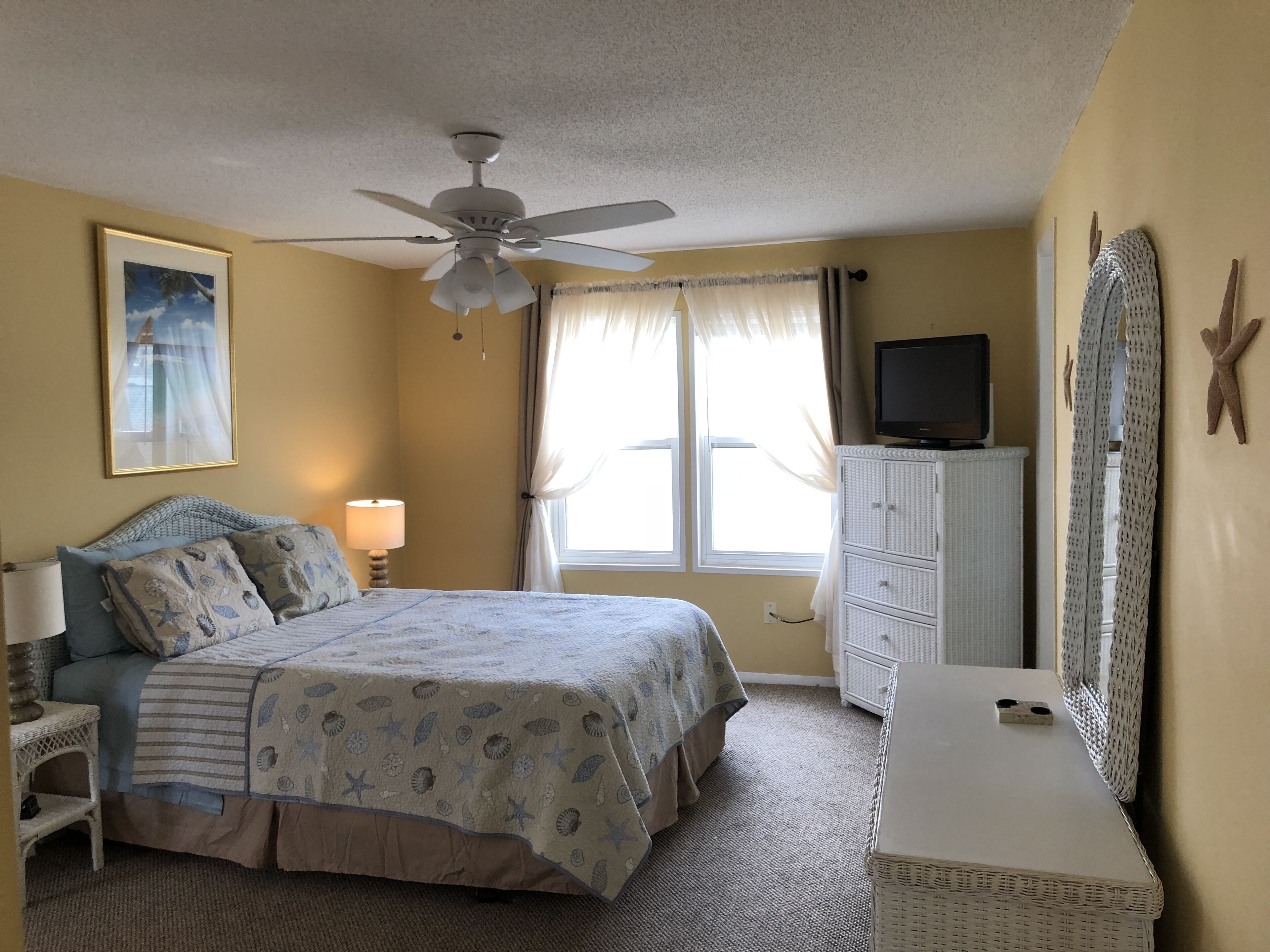 A bedroom with a bed, a ceiling fan, a television, a dresser, and a window with curtains.