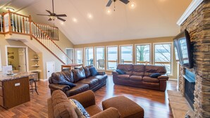 A spacious living room with a high ceiling, wooden staircase, and large windows offering a view of the outdoors. - Bridgeview Cottage with Weekday Pontoon Boat Access, and Quiet Peninsula Setting (Cadiz)