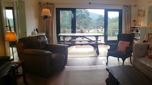 Una sala de estar con un sofá, dos sillones, una mesa de centro de madera y una mesa de comedor con sillas. Hay un ventanal con vistas a árboles y montañas.