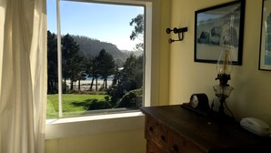 Habitación con un ventanal grande que ofrece vista a una playa y colinas. Hay un aparador de madera con una lámpara y un reloj encima.