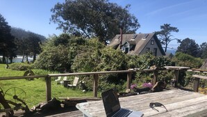 Una terraza de madera con una laptop sobre un banco, con vista a un jardín con sillas y una casa al fondo.