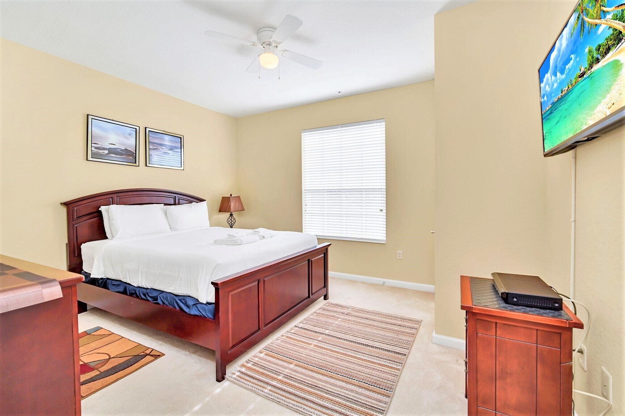 A bedroom with a bed, a ceiling fan, a TV mounted on the wall, and a window with blinds.