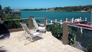 A terrace with lounge chairs overlooking a body of water, a dock with boats, and a distant island with buildings. - Luxury Waterfront Condo w/ Pool,  Dock, and Gardens  (West End)
