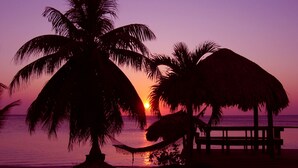 A beach scene with a sunset, palm trees, and a thatched-roof structure. - Luxury Waterfront Condo w/ Pool,  Dock, and Gardens  (West End)