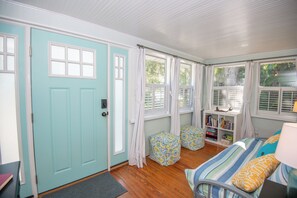 A room with a teal door, wooden flooring, a bed with striped bedding, two patterned ottomans, a bookshelf, and a window with curtains.