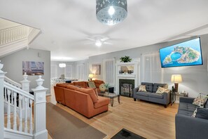 A spacious living room with a staircase, a fireplace, and a television displaying a scenic view. - Beach Block SHORE HOUSE  7BR Sleeps 16 Hot Tub! Ocean Views & Steps to Boardwalk (Atlantic City)