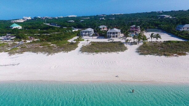 Une vue aérienne d’une plage aux eaux turquoise et limpides, avec un rivage sablonneux et plusieurs bâtiments dotés de grandes fenêtres et de balcons.