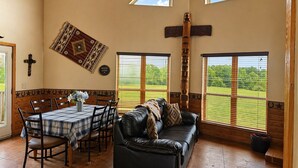 A room with a dining table, chairs, a leather sofa, a cross on the wall, a wooden totem, and large windows with a view of a green field.
