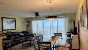 A living room with a sofa, a dining table with chairs, a ceiling fan, and a large window. - Oceanfront! Heated Pool! Free WiFi & Cable! Bidets! Deluxe Outdoor Kitchen! Oceanway 203! (Indian Rocks Beach)
