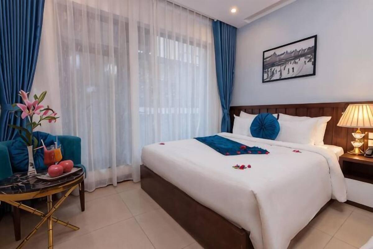 A hotel room with a large bed, a bedside table with a lamp, a small round table with a vase and a drink, and a window with sheer curtains.