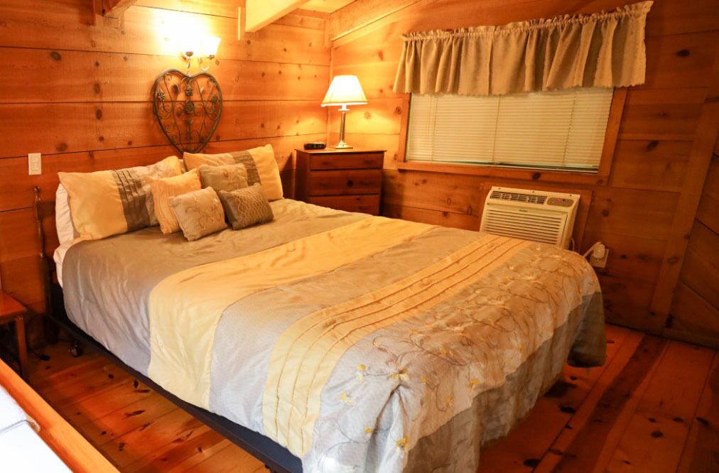 A cozy wooden cabin bedroom with a large bed, a nightstand with a lamp, a heater, and a window with curtains.