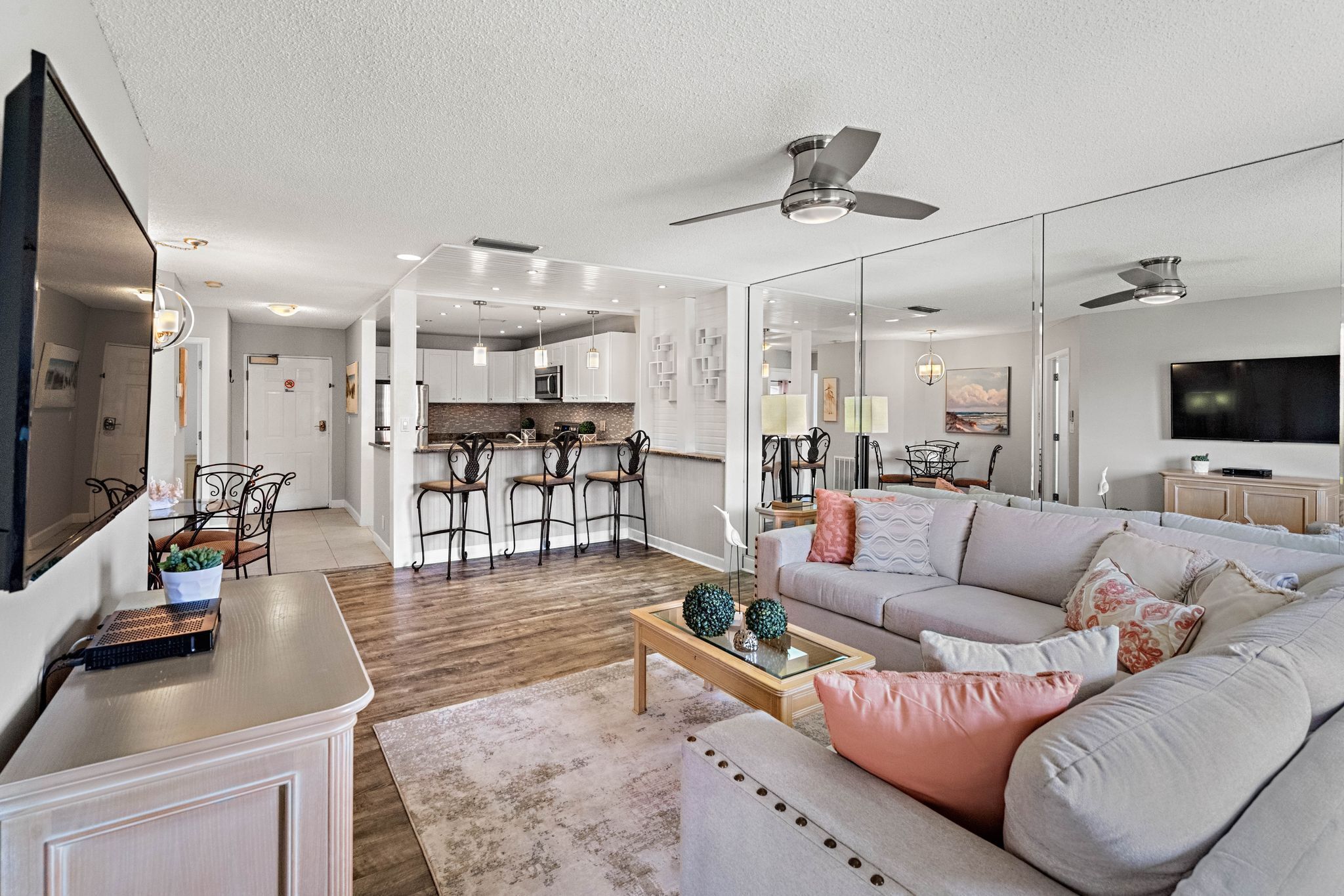 A spacious living room with a large sofa, a coffee table, and a TV. There is a kitchen in the background with a dining area.