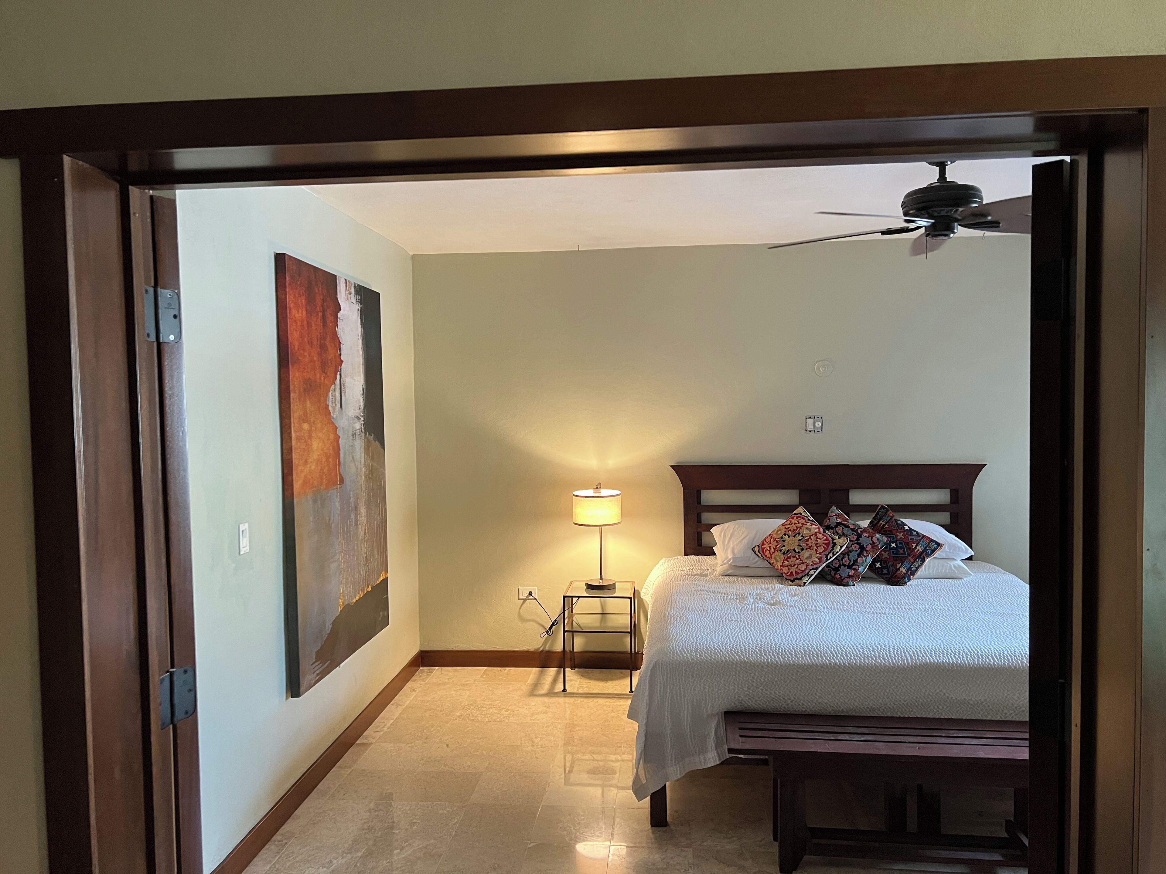 A bedroom with a bed, a ceiling fan, a nightstand, a lamp, and a painting on the wall.