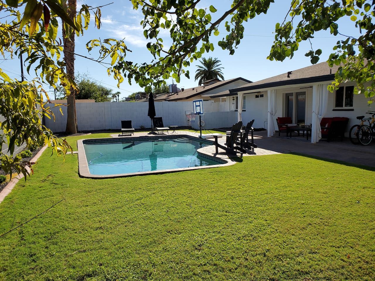 En have med swimmingpool, en terrasse med stole og et bord, samt en hvid bygning med dør og vinduer.