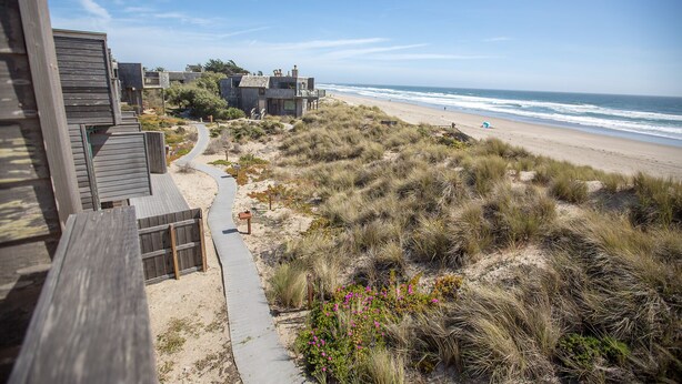 Una propiedad costera con una terraza de madera, un sendero arenoso y vista a la playa y al océano.