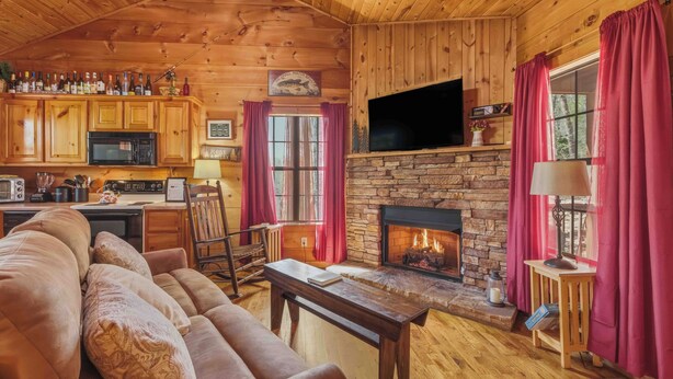 A cozy cabin interior with a stone fireplace, wooden furniture, a kitchen area, and a television mounted above the fireplace. - Couples Retreat - Romantic and Relaxing Cabin Retreat (Cleveland)
