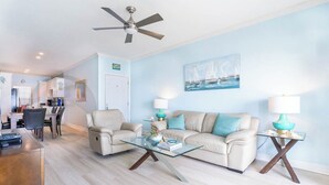 Una sala de estar con una mesa de centro de vidrio, dos sillones, un sofá y un comedor con una cocina al fondo.