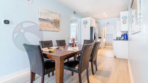 Un comedor con una mesa de madera y sillas negras, una cocina con armarios blancos y una pared azul claro con un cuadro de temática marina.