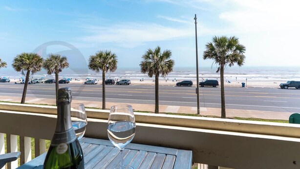Un balcón con vista a la playa y al océano, con palmeras y coches aparcados.