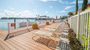 En träterrass med solstolar och krukväxter, med utsikt över en småbåtshamn där båtar och byggnader syns i bakgrunden.