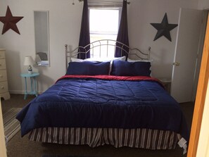 Une chambre à coucher avec un cadre de lit en métal, une couvre-lit bleue et rouge, un couvre-lit à rayures, une petite table de chevet bleue avec une lampe, un miroir et deux étoiles peintes sur les murs.