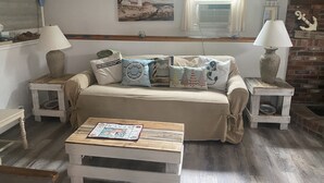 Un salon chaleureux avec un canapé beige, deux tables d’appoint avec des lampes, une table basse et un tableau représentant un phare.