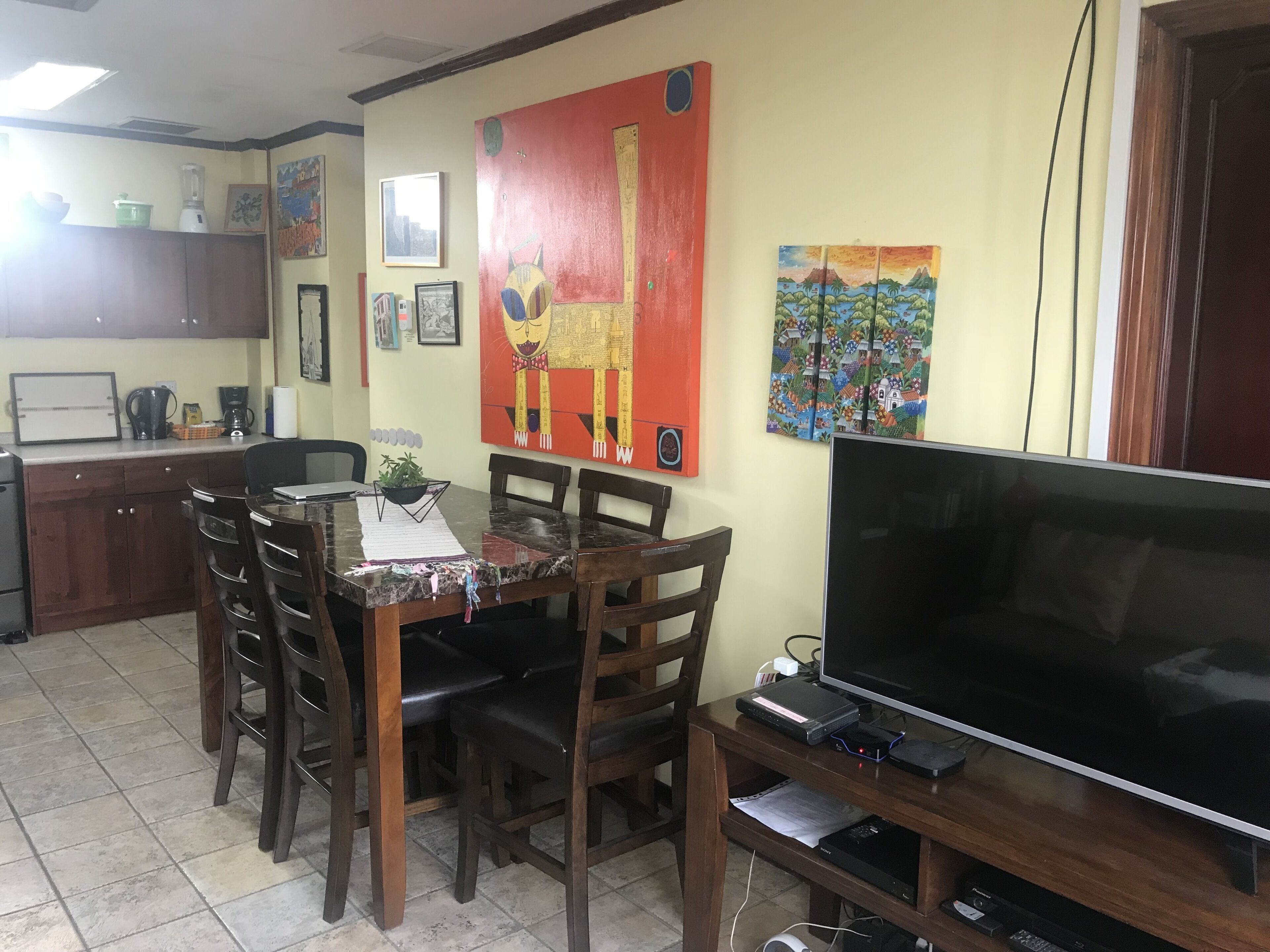 A dining area with a table, chairs, a TV, and a kitchen in the background.