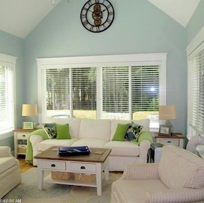 Relax in this spacious living room with a sofa, table, and clock, surrounded by natural light and a high ceiling.