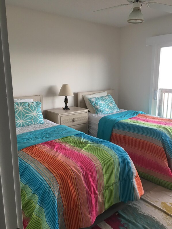 A bedroom with two beds, each with a colorful striped bedspread. There is a nightstand with a lamp and a drawer between the beds. - Queen's Grant Oceanfront Condo, Topsail Beach, 1st Floor, Pool, Boat access. (Topsail Beach)