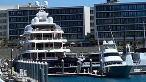 A marina with several boats docked, a large building with multiple floors in the background, and a clear blue sky.