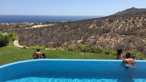 A swimming pool with clear blue water, surrounded by a grassy area and a view of a desert landscape with a mountain in the distance.