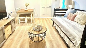 Un espace de vie compact comprenant un coin repas, un canapé agrémenté de coussins décoratifs, une table basse ronde et un téléviseur posé sur un meuble en bois.