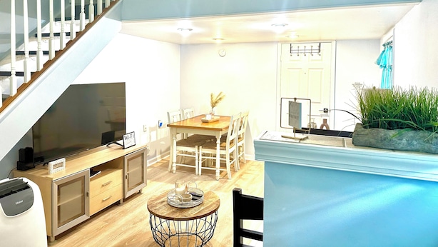Un vaste espace de vie comprenant un escalier en bois, une table à manger avec des chaises, un téléviseur et un coin cuisine équipé de placards et d’une machine à café.