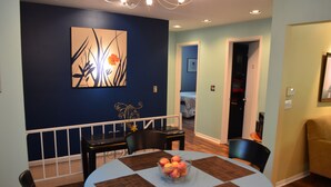A dining area with a round table, chairs, and a bowl of fruit. There is a painting on the wall and a door leading to another room.