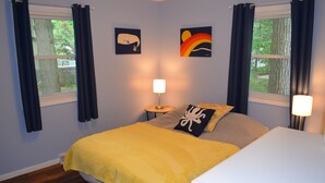 A bedroom with a bed, two bedside lamps, a window with curtains, and two wall art pieces.